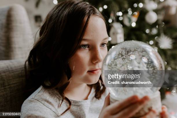 girl sitting by a christmas tree looking at a snow globe - bola de cristal com neve imagens e fotografias de stock