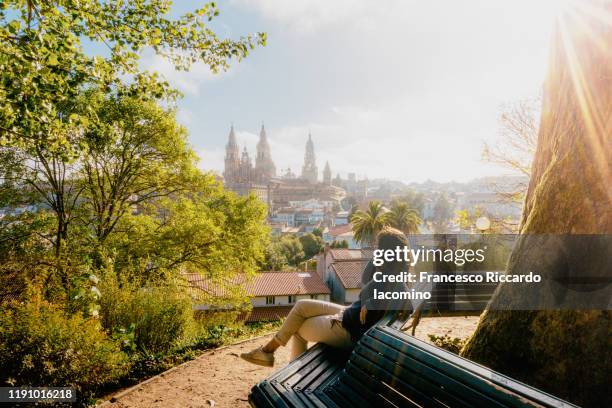 woman watching santiago de compostela cathedral at sunrise, park and sunny sky. galicia, spain - bench park stock-fotos und bilder