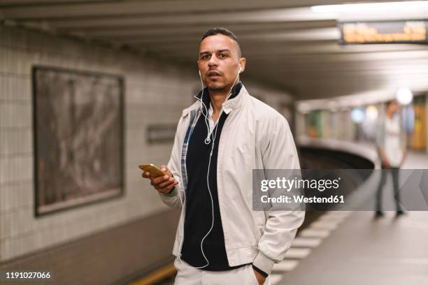 portrait of man waiting at underground station platform listening music with earphones and cell phone - metro platform stockfoto's en -beelden