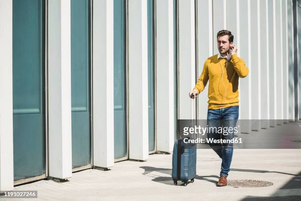 young man with rolling suitcase and earphones in the city on the go - yellow suitcase stock pictures, royalty-free photos & images