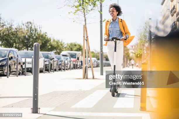 happy young woman with earphones riding e-scooter in the city - tretroller stock-fotos und bilder