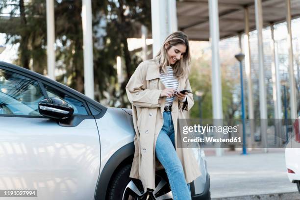 young blond woman using smartphone, leaning on a car - trench coat ストックフォトと画像