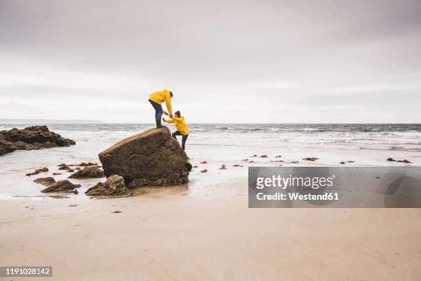 young woman wearing yellow rain jackets and climbing on a rock at the beach, bretagne, france - bretagne photos et images de collection