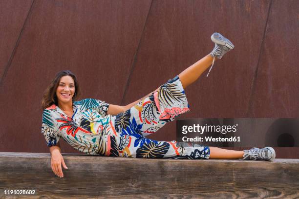 portrait of smiling young woman lying on wooden bench lifting leg - multi coloured trousers stock pictures, royalty-free photos & images