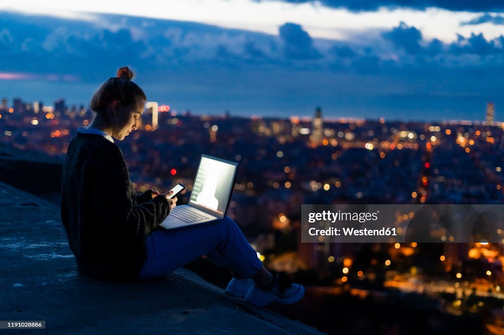 Young woman using cell phone and laptop at dawn above the city, Barcelona, Spain