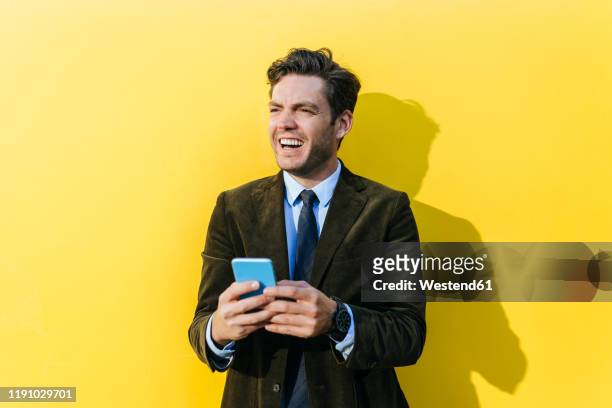 happy businessman with smartphone in front of yellow wall - brown suit stock pictures, royalty-free photos & images