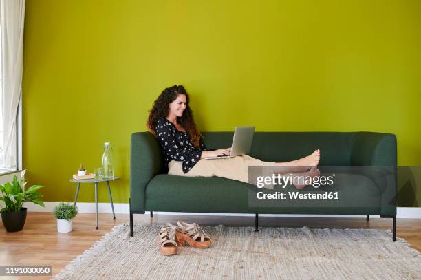 smiling young businesswoman using laptop on couch in modern office - coloured wall stockfoto's en -beelden