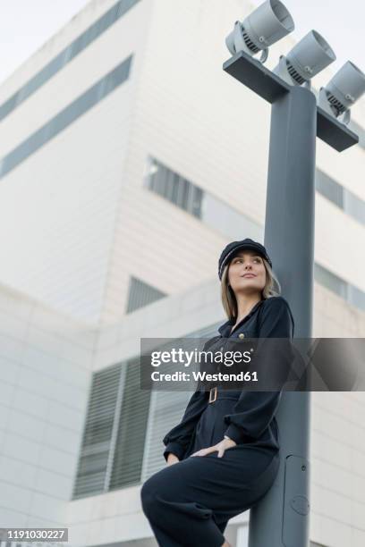 young blond businesswoman wearing black sailor's cap and looking sideways, leaning on a lamp pole, low angle view - poteau dappui photos et images de collection