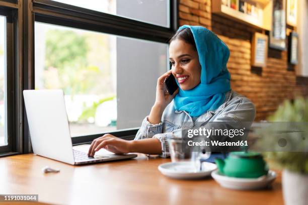 businesswoman wearing turquoise hijab in a cafe and working with her laptop, on the phone - arabic keyboard fotografías e imágenes de stock