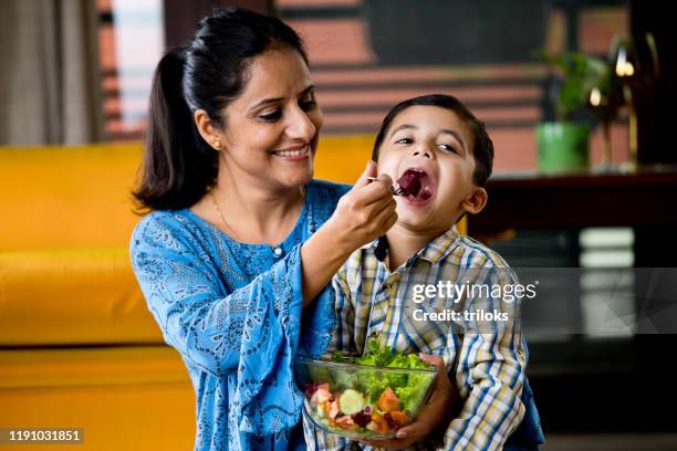happy mother feeding fruit salad to son - indian mother and child stock pictures, royalty-free photos & images