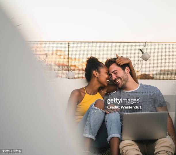 happy young couple with laptop and earphones sitting on rooftop in the evening - summer nights stock-fotos und bilder