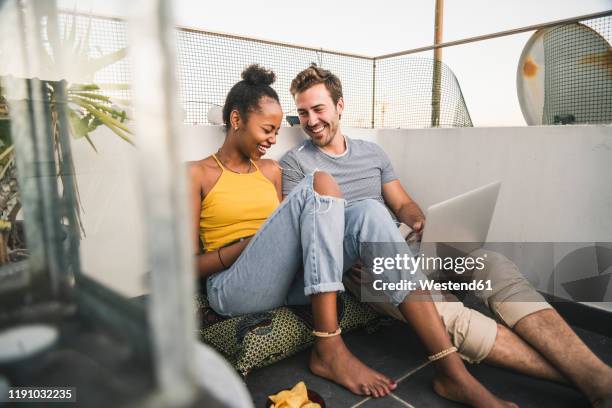 happy young couple with laptop and earphones sitting on rooftop in the evening - couple balcony stock-fotos und bilder