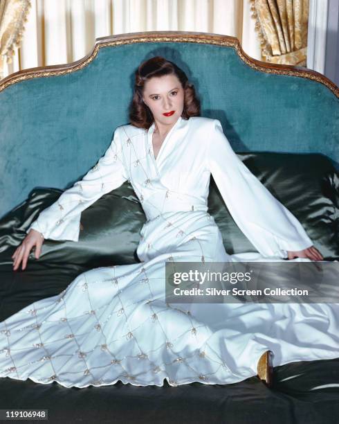 Barbara Stanwyck , US actress, wearing a long white dress, reclining on a bed with silk bedding, circa 1940.