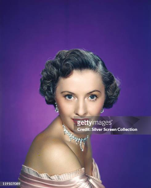 Headshot of Barbara Stanwyck , US actress, poses wearing a shoulderless dress with diamond earrings and necklace in a studio portrait, against a...