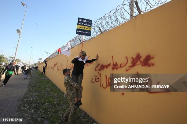 Member of an Iraqi Shiite paramilitary group helps a fellow protesters holding a placard denouncing the United States climb on the outer wall of the...