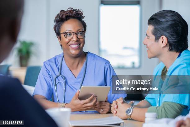 female nurse holding digital tablet while sitting at conference table with colleagues - resident stock pictures, royalty-free photos & images