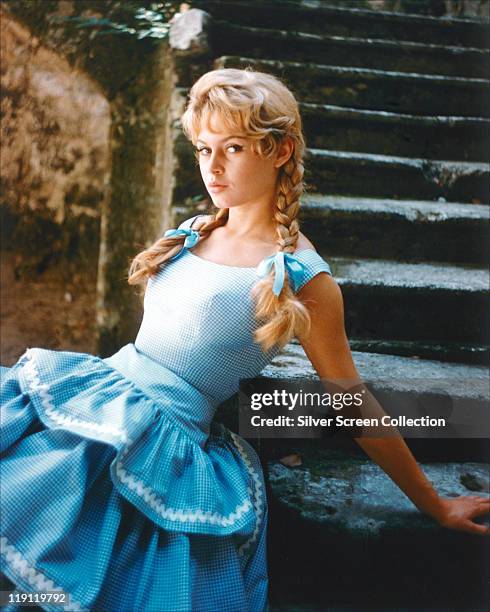 Brigitte Bardot, French actress, model and singer, wearing a blue gingham dress with her hair in plaits, tied with blue bows, sitting in a series of...