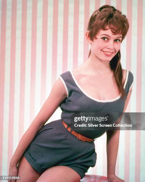 Brigitte Bardot, French actress, model and singer, wearing a blue playsuit, with a red belt, in a studio portrait, against a pink-and-white striped...