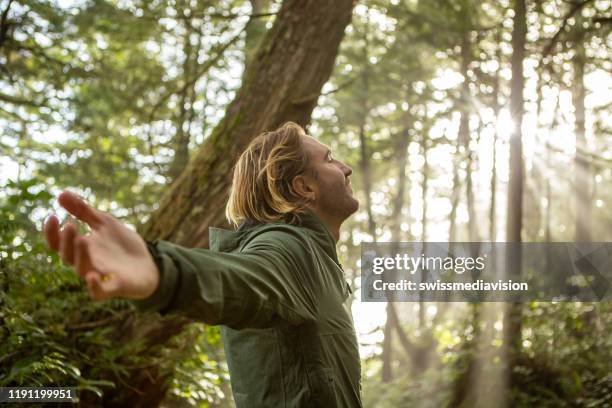 jeune homme embrassant la forêt tropicale restant dans les rayons du soleil illuminant les arbres - les bras écartés photos et images de collection