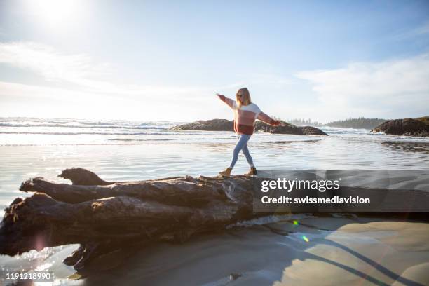 young woman walking on tree trunk on beach balancing body weight - british columbia beach stock pictures, royalty-free photos & images