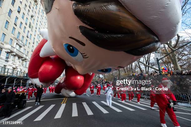 Balloon handlers fight the strong wind gusts and struggle but Santa's Scout Elf, The Elf on the Shelf Balloon leg hits the ground as he makes his way...