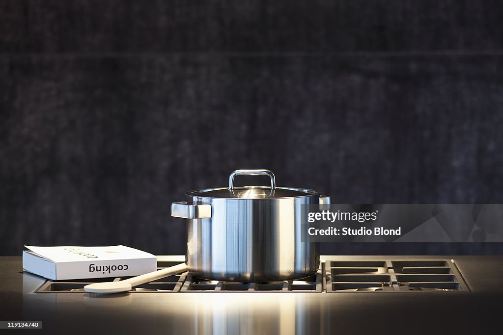 Saucepan, cookery book and wooden spoon on hob.