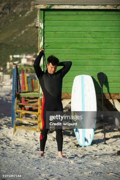 male surfer with paddle surfboard pulling wetsuit zip - paddle board men imagens e fotografias de stock