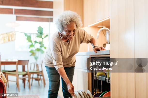 mulher sênior de sorriso que mantem placas na máquina de lavar louça - bending - fotografias e filmes do acervo