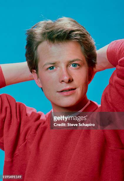 Portrait of Canadian actor Michael J Fox, in a red sweater, as he poses with his hands behind his head, against a blue background, Los Angeles,...
