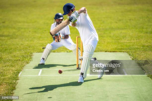 cricket batter hitting the ball while the wicket-keeper stands behind him. - batsman stock pictures, royalty-free photos & images