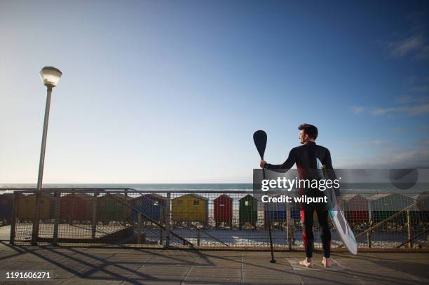 full-length man with wetsuit standing with stand up paddleboard overlooking colourful beach huts - midlife crisis stock pictures, royalty-free photos & images