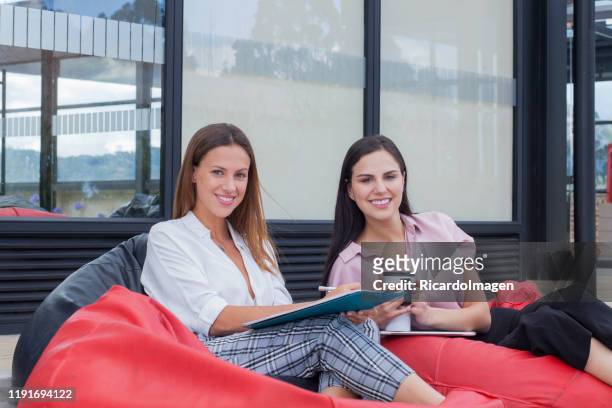 lateinische mitarbeiterinnen sitzen in rotem pouf auf der terrasse der firma, für die sie arbeiten, sie schauen in die kamera und lächeln - office smile light stock-fotos und bilder