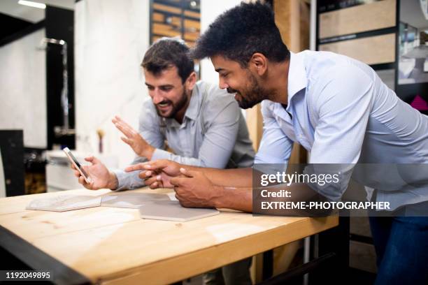 two men leaning over the desk covered in hexagonal wooden tiles, looking together at a mobile phone in a shop - customer experience stock pictures, royalty-free photos & images