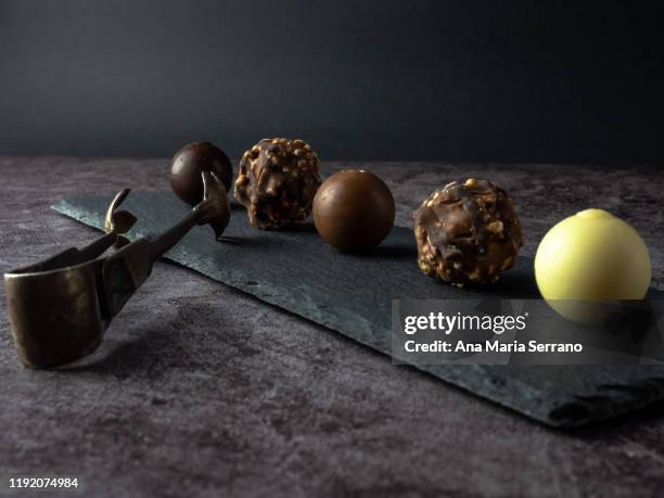 homemade chocolate crunchy chocolates with hazelnut and dried fruit pieces, white chocolate bonbons, dark chocolate and brown chocolate on an elongated slate plate with an old alpaca serving tongs on a dark background - pastry imagens e fotografias de stock