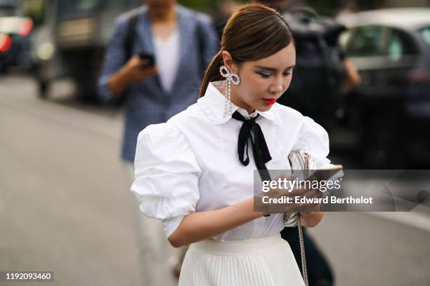 Guest wears earrings, a white blouse with a frilly collar, puff sleeves, and a black lavaliere, a shiny silver-tone Balenciaga bag, a cream-color...