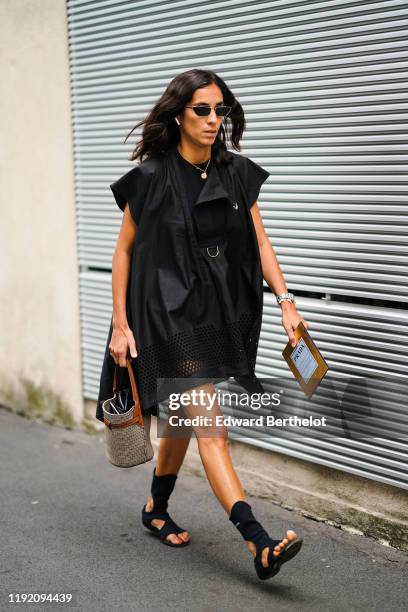 Guest wears sunglasses, a black oversized flowing mini dress, a brown and black houndstooth bucket bag, black Prada gladiator flat sandals, outside...