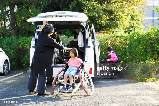 young girl sitting on her sister’s tennis wheelchair with her sister in a wheelchair in the back and their mother closing the rear door of the car - japanese tennis stock-fotos und bilder