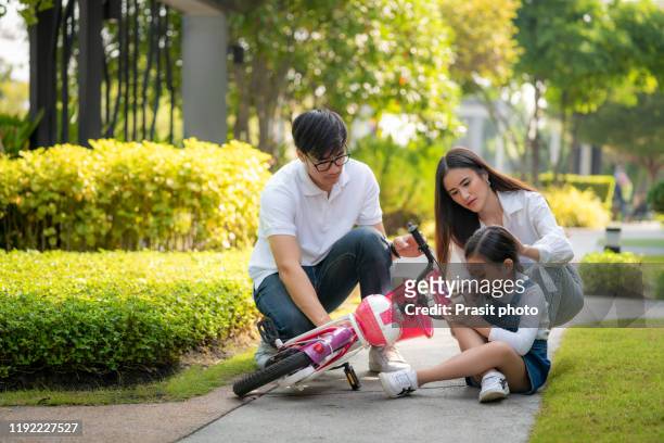 asian family with father and mother helping and checking on leg her daughter has accident while practice to riding a bicycle at park in village. - asian family fall stock-fotos und bilder
