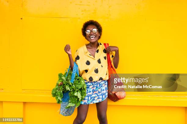 one young woman carrying grocery bags in front of yellow wall laughing - fashion food stock pictures, royalty-free photos & images