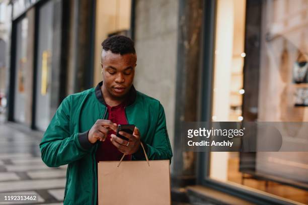 handsome african man using smartphone while holding shopping bags - chubby man shopping stock pictures, royalty-free photos & images