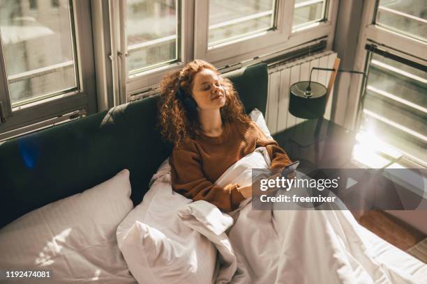 young woman sitting on bed and using mobile phone - sunny window stock pictures, royalty-free photos & images