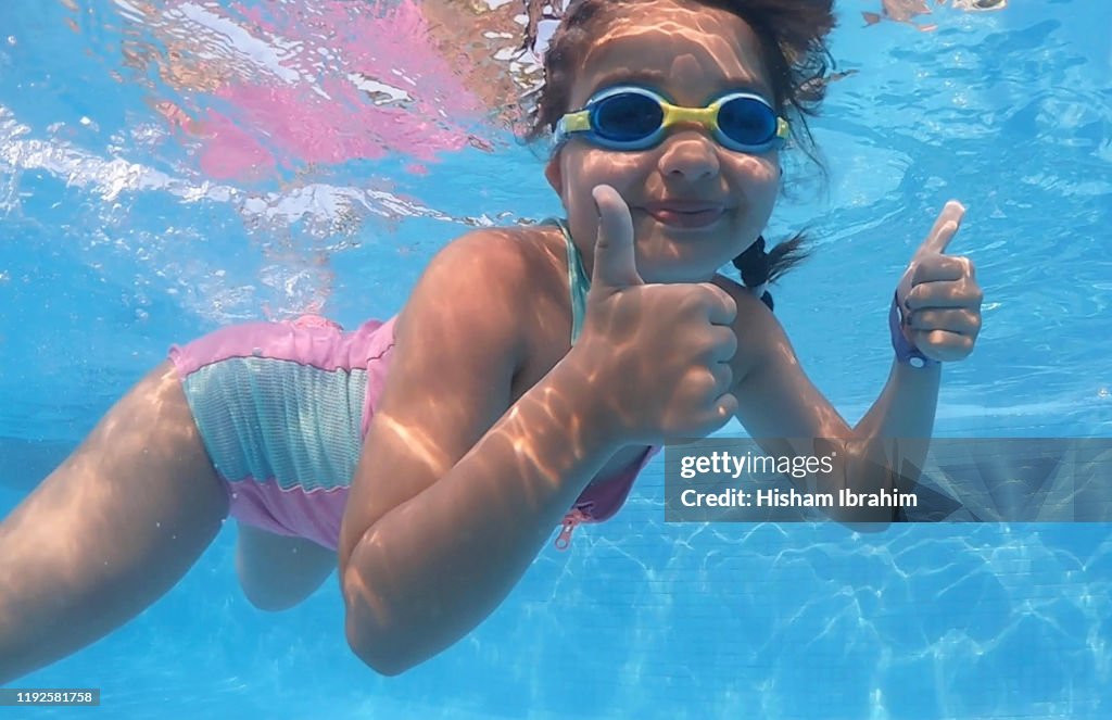 7 years old girl swimming underwater in swimming pool showing thumbs up.