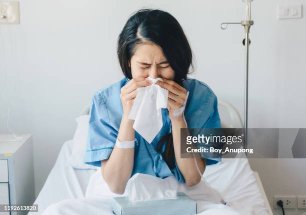portrait of young woman wearing patient cloths sneezing in tissue at hospital. - mouchoir photos et images de collection