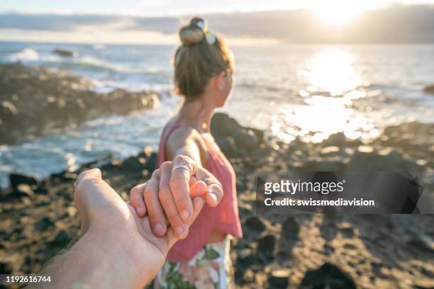 couple holding hands on rocky shoreline at sunrise, follow me - follow me to man stock pictures, royalty-free photos & images
