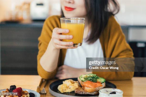 smiling young asian woman holding a glass of juice while having breakfast at home - woman drinking juice stock pictures, royalty-free photos & images