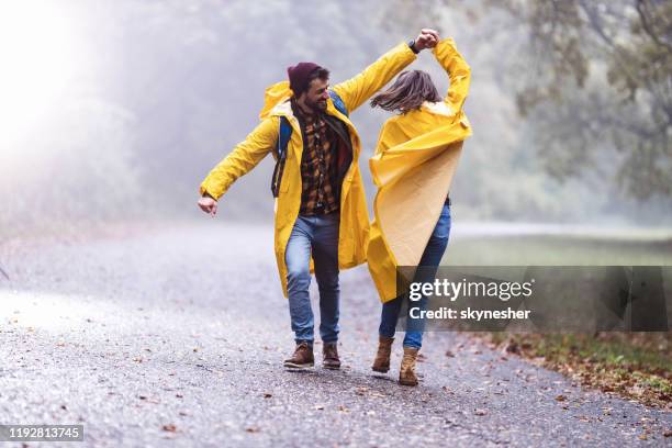 glückliches paar in regenmänteln, die spaß beim tanzen auf einem regen im n:0 haben. - winter friends playing stock-fotos und bilder