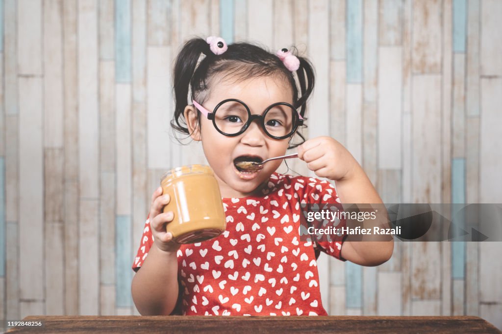 Portrait Of Girl Eating Peanut Butter At Table Against Wall
