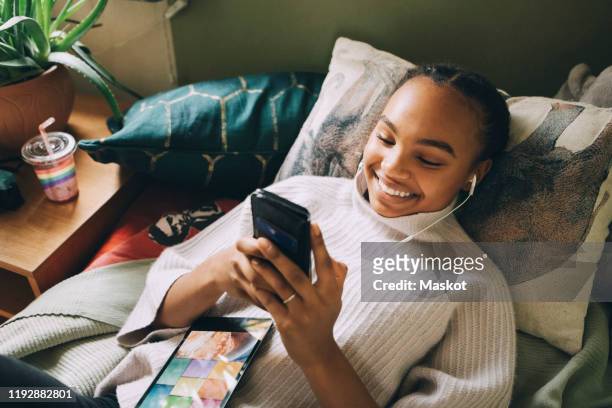 high angle view of smiling teenage girl listening music while using mobile phone on bed at home - listening stockfoto's en -beelden
