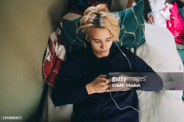 high angle view of male teenager text messaging on mobile phone while lying in bedroom - boy teenager stock-fotos und bilder