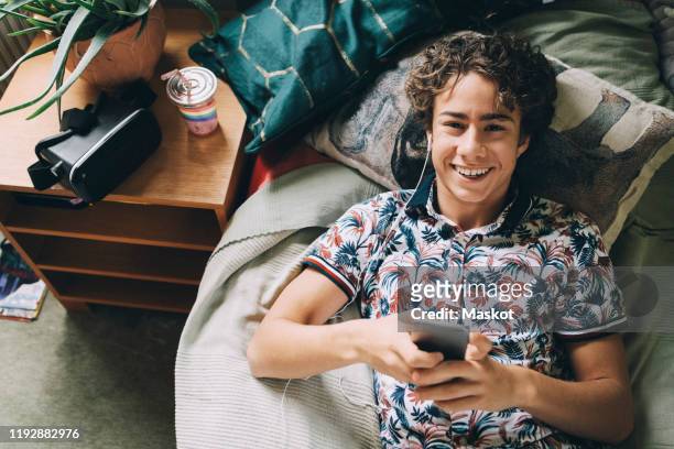 high angle portrait of happy teenage boy listening music through smart phone on bed at home - one teenage boy only stock pictures, royalty-free photos & images